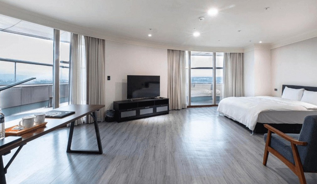 新舍商旅中壢館的套房，設計以淡色調為主，中央擺放大床，旁邊有獨立的桌椅和沙發區，室內電視與寬大落地窗使空間更顯通透，窗外陽台可眺望遠景，適合長時間入住或家庭旅遊使用。