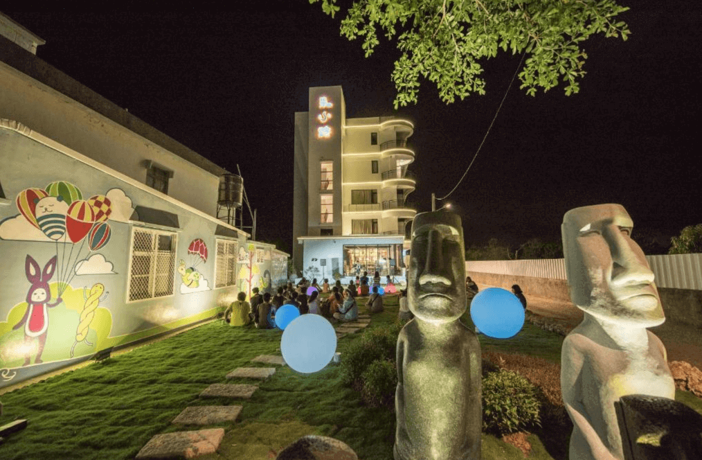 【小琉球住宿】朵小路北歐民宿的夜晚戶外庭院，草坪上擺放著摩艾石像裝飾，並配有彩色燈光，建築外牆繪有可愛的卡通圖案，展現歡樂的氣氛。