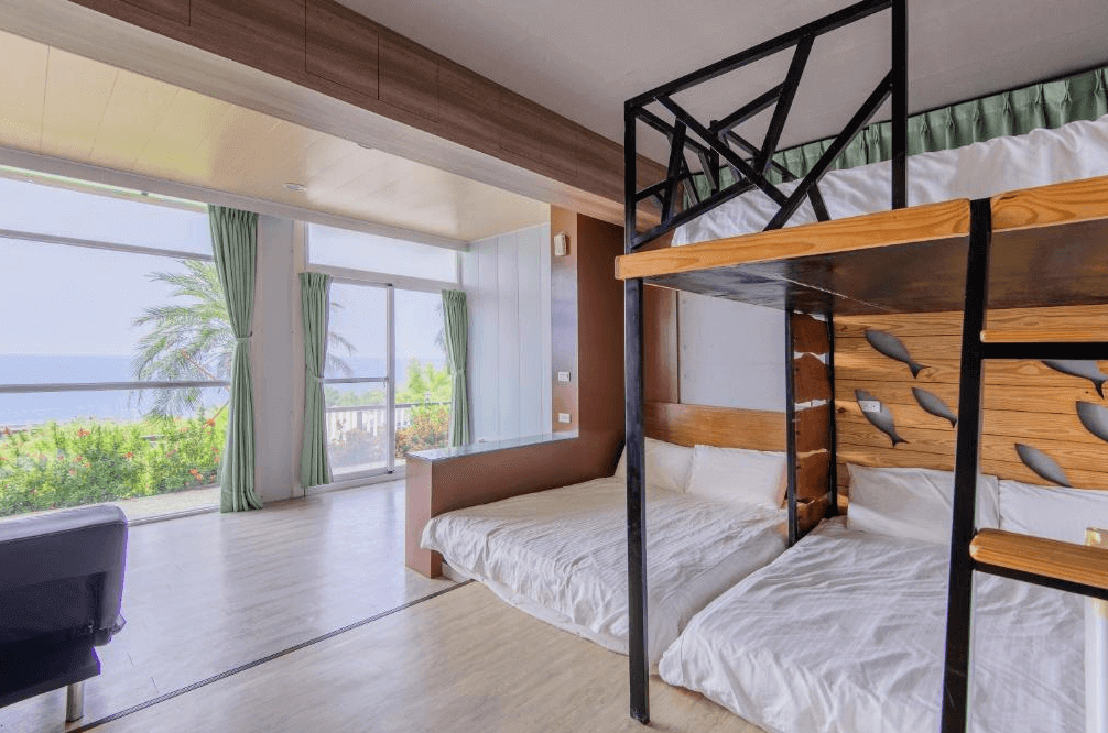 【小琉球住宿】杉福海灣渡假會館的家庭房設計，內有一組上下鋪木床與地板床，牆面裝飾有魚群圖案，窗外景色是充滿熱帶風情的綠樹與海洋，展現舒適的家庭氛圍。