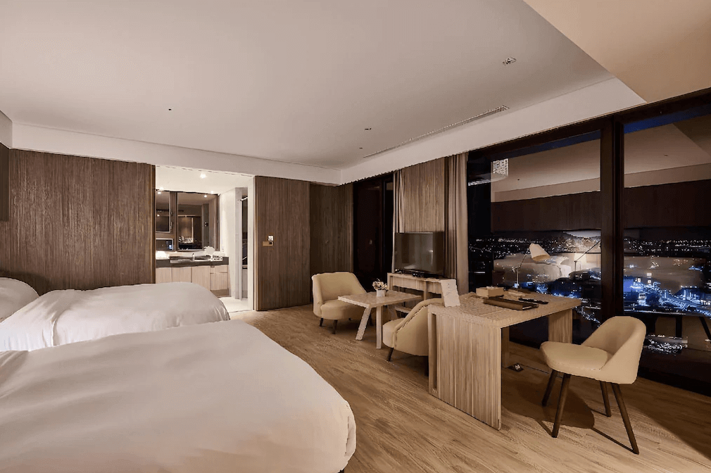 村却國際溫泉酒店的寬敞雙床房，內裝以溫暖的木質調為主，擁有大面積落地窗，可以欣賞夜景。房間內設有簡約的書桌、沙發椅，並且有開放式的設計，透過門可以看到浴室的部分。