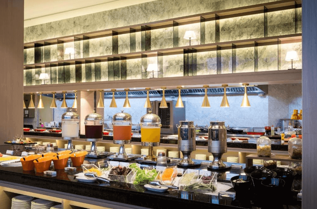 村却國際溫泉酒店的豐富自助早餐吧檯，擺放著各式果汁和沙拉材料。裝潢以大理石和木質為主，燈光溫馨，並且有金屬材質的飲品保溫壺，營造出高雅的用餐氛圍。