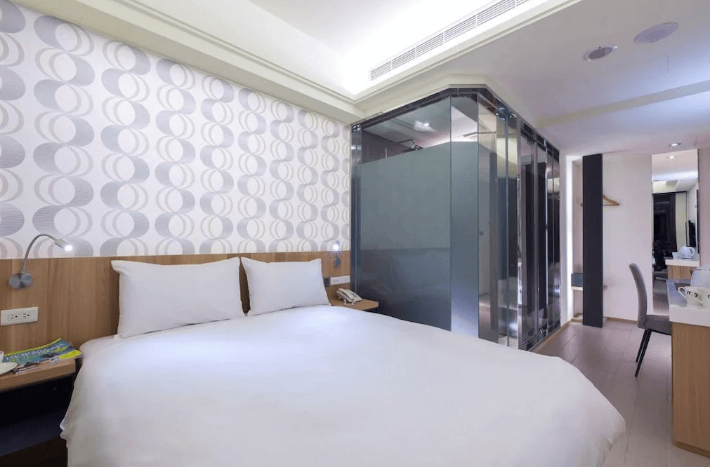 【中壢火車站住宿】“中壢夜市住宿”沐楓商旅的客房，床頭牆面採用幾何圖案裝飾，床旁配置了閱讀燈和小型床頭櫃，浴室以透明玻璃隔間與房間分離，空間寬敞明亮。