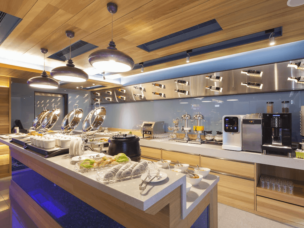 【中壢火車站住宿】“中壢夜市住宿”沐楓商旅酒店餐廳的自助早餐區，設計以木質為主，吊燈營造溫暖氛圍，餐檯上擺放多種餐點，包括麵包、湯品和飲品，設備齊全，展現豐富的用餐選擇。