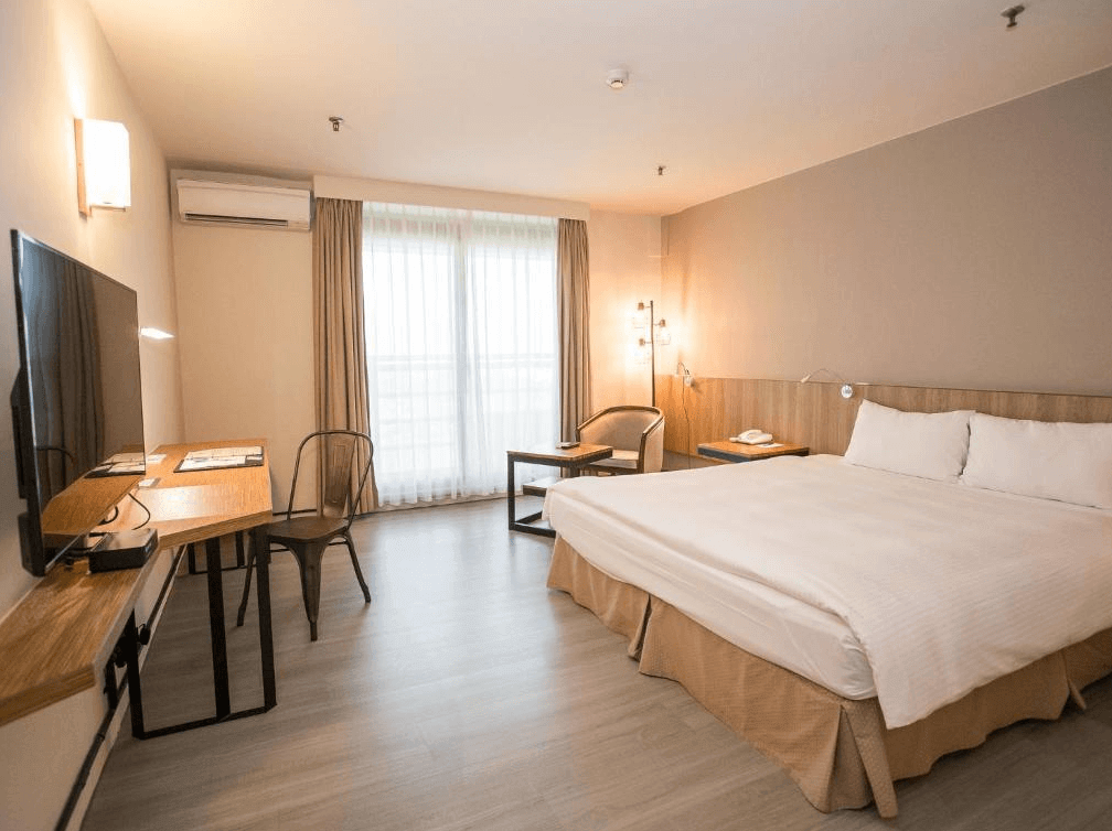 玖屋文旅配有大床的簡約雙人房，採用暖色調裝潢，房間內設有一張木質書桌和金屬椅，窗戶透進自然光線，營造出舒適的住宿環境。