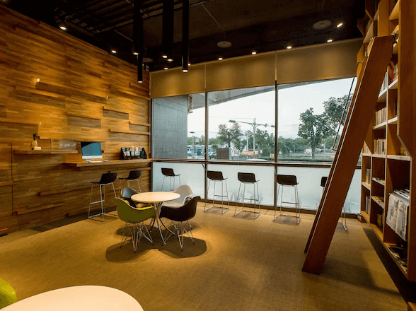 玖屋文旅的舒適閱讀休憩區域，設有現代感十足的木質牆面和大面積玻璃窗。空間內有多張圓桌椅，並有一個書架，適合用來閱讀或輕鬆放鬆。