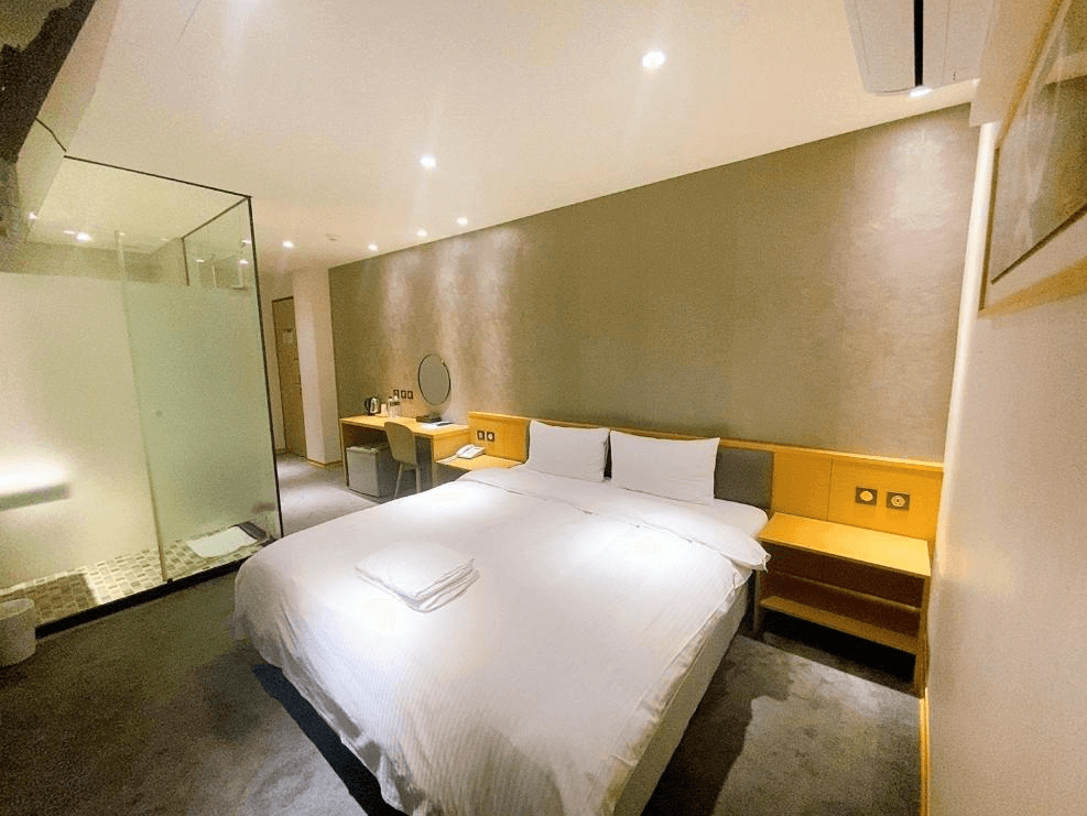 【中壢火車站住宿】“中壢夜市住宿”米淇旅店中壢館的雙人房，配置一張大床，房內有書桌、化妝鏡和座椅，浴室與臥室以透明玻璃隔開，整體設計簡潔實用，燈光柔和營造溫馨感。