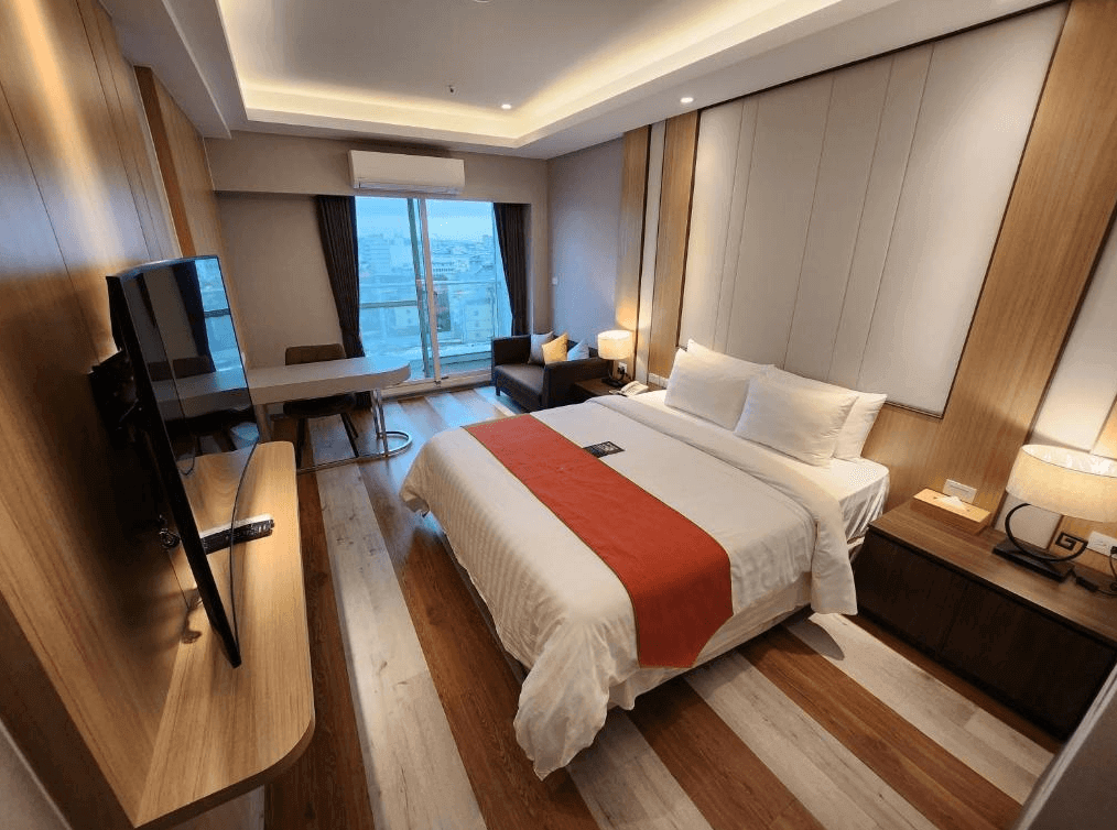 賓利酒店的客房，配有木質地板和牆面，中央擺放一張鋪有白色床單和橘色條紋毯的雙人床，房間內設有辦公桌、沙發和落地窗，窗外可見城市景色，整體設計溫暖且舒適。