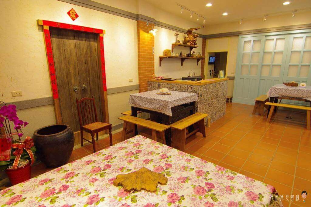 【北港住宿】北港好住背包客棧的餐廳區域以木質餐桌椅和花卉桌布為主，搭配紅磚牆與藍色木門，增添了傳統與懷舊感。牆上擺放著裝飾品，營造出溫馨且家的氛圍。