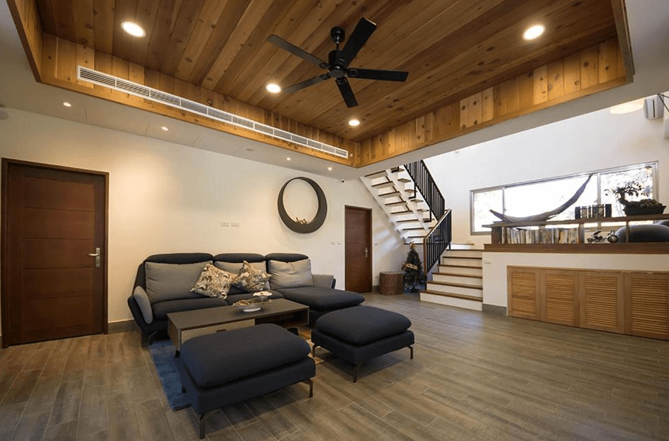 【通霄住宿】南窩綠丘的開放式現代客廳，木質天花板搭配吊扇，空間寬敞明亮。中央擺放著一組深藍色沙發及茶几，後方樓梯通往二樓，整體設計充滿自然質感且功能齊全。