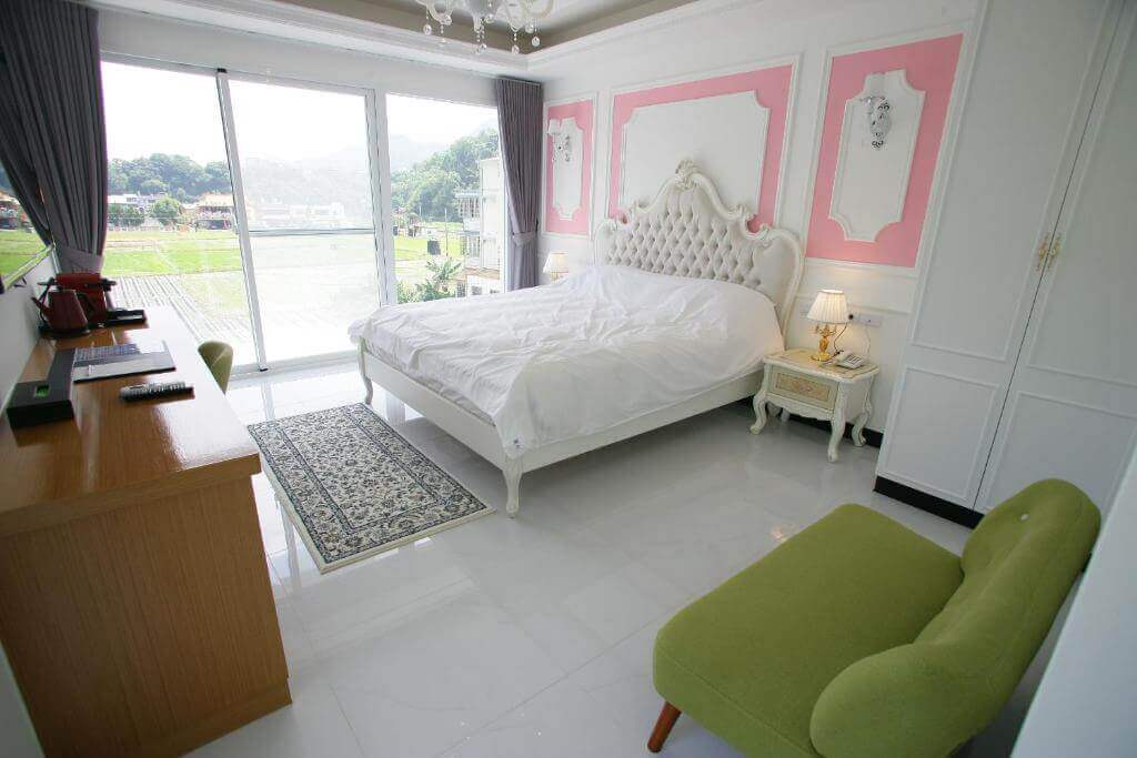 【大湖住宿】夏內爾落羽松民宿粉色與白色相間的牆面設計，搭配雕花床頭板與金色台燈，營造出浪漫且溫馨的氛圍，大面落地窗提供充足的自然光並享有田園景色，整體空間明亮寬敞。