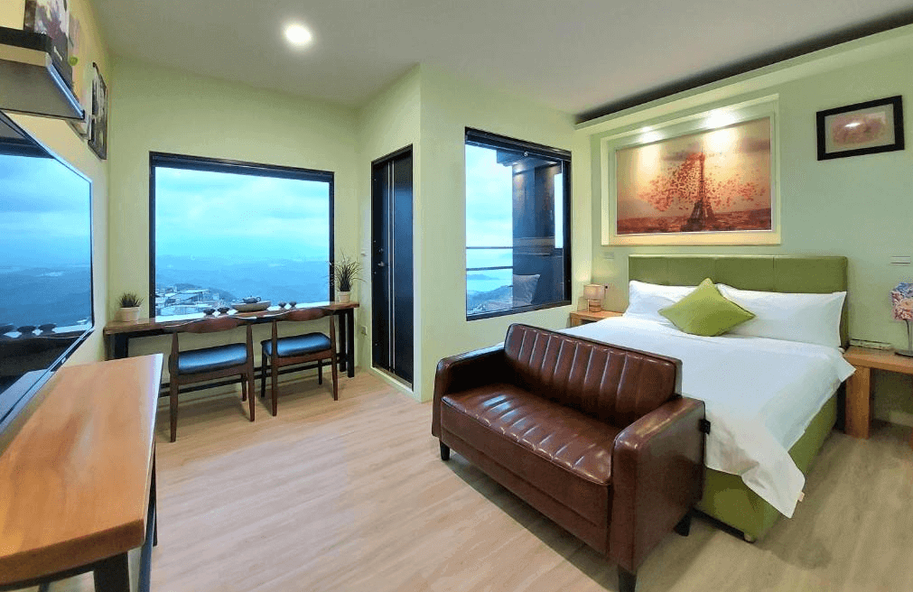 【九份寵物友善住宿】多桑的店民宿的房間內設有一張大床，搭配綠色床頭板與白色床品，營造出清新舒適的氛圍。床前擺放著一張棕色皮沙發，增添居家感。房間內設有大片窗戶與書桌，能欣賞窗外山景與海景，光線明亮且視野遼闊。