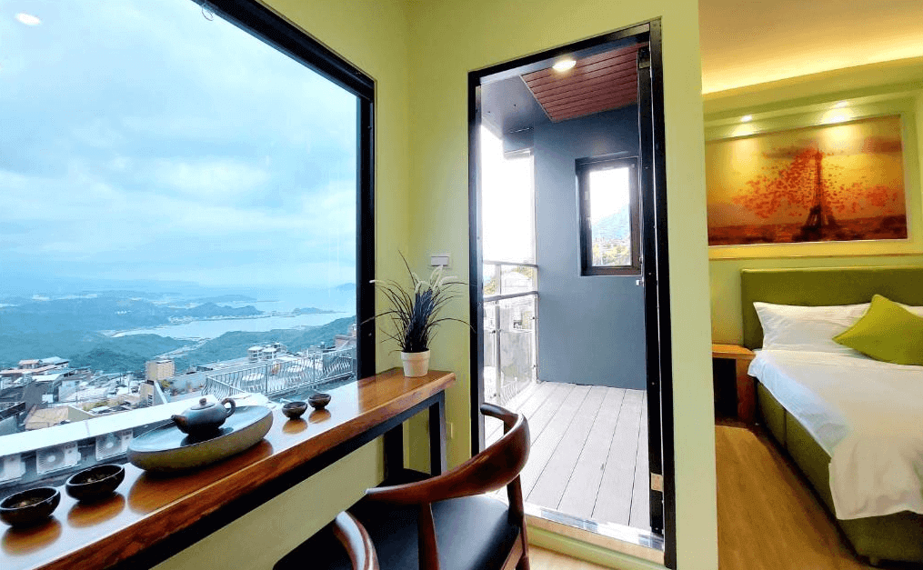 【九份寵物友善住宿】多桑的店民宿近距離展示窗外的景觀，綠意山巒與海灣一覽無遺。房間內的小陽台與開闊的玻璃窗設計，讓旅客能隨時欣賞美景。桌上擺放簡約茶具與綠植，增添一絲文藝氣息。