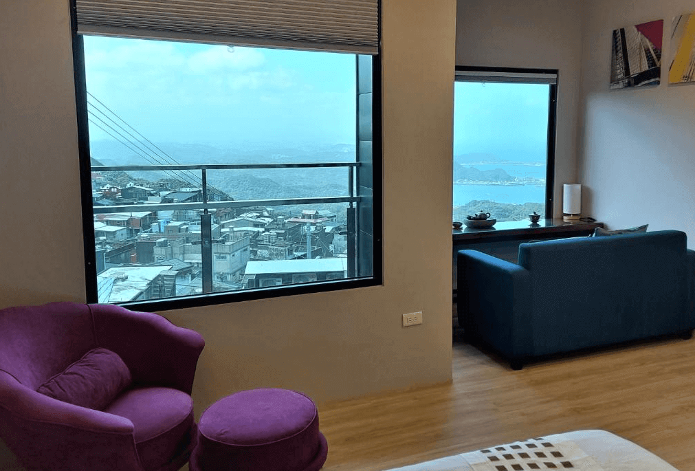 【九份寵物友善住宿】多桑的店民宿的房內不同角度景觀視角，擺放一張紫色休閒椅與腳凳，為旅人提供放鬆休憩的角落。另一面窗戶則能看到層疊的山城建築與壯麗海景，室內搭配深藍色沙發，整體風格優雅舒適。