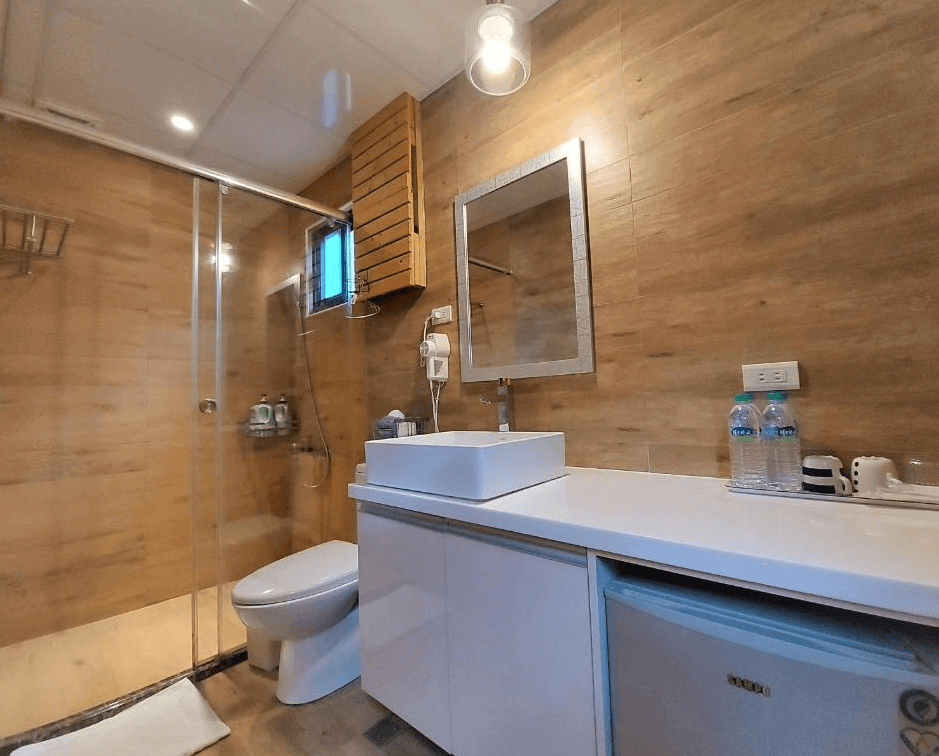 【九份寵物友善住宿】多桑的店民宿的浴室設計簡約現代，牆面與地板選用木質紋理風格，營造自然溫暖感。設有乾濕分離的玻璃淋浴間、乾淨整潔的洗手檯，並配備瓶裝水、吹風機等基本設施，滿足旅客需求。