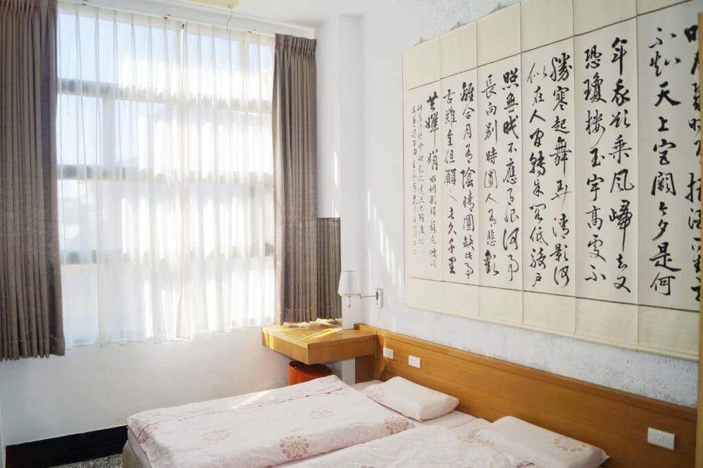 【北港住宿】好住民宿的房間以傳統中式書法掛畫為裝飾，搭配木質床頭與淺粉色床單，展現濃厚的文化氛圍，同時窗戶引入充足光線，讓空間更顯寬敞明亮。