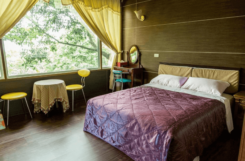 【通霄住宿】如意園民宿內部的雙人房設計，木質牆面搭配深紫色床單，床邊設有小型梳妝台。大片窗戶外樹影婆娑，自然光灑入，營造舒適溫馨的氛圍，適合放鬆休息。