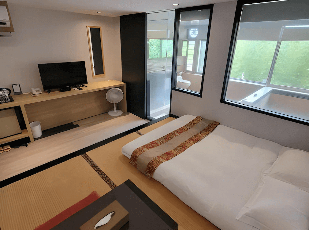 【烏來住宿】山水妍溫泉會館的日式榻榻米房間，內部裝潢以簡約風為主，配有落地窗和自然採光，室內設施包括電視和書桌，浴室區域透明玻璃設計，窗外綠意盎然。