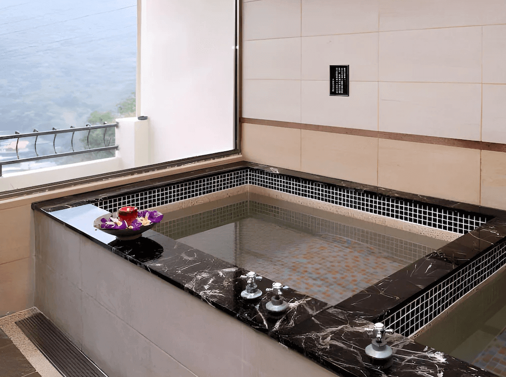 【烏來住宿】山水妍溫泉會館的室內溫泉池設計高雅，以大理石台面和淺色瓷磚搭配，池邊裝飾著花卉，透過窗戶可欣賞戶外青山景色，氛圍寧靜放鬆。