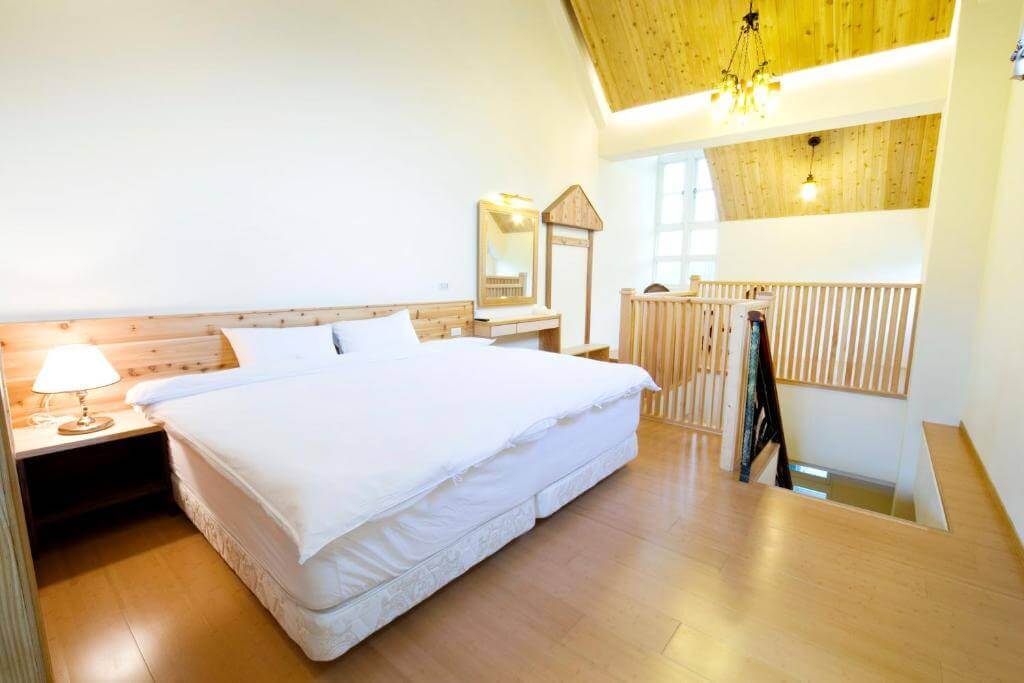 【大湖住宿】巧克力雲莊民宿的雙人房，採用溫暖的木質調設計，搭配高挑天花板與優雅吊燈，讓空間顯得舒適且通透，並提供基本的床邊燈與化妝鏡，符合旅客對便利與舒適的需求。