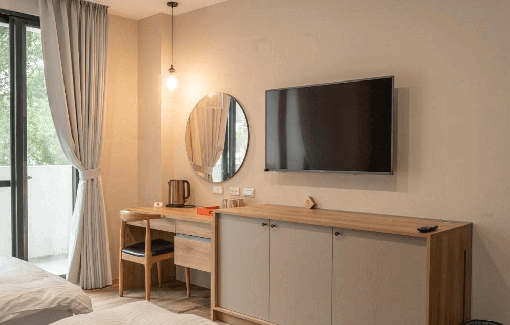 【水里住宿】恩典花園咖啡民宿臥室內的電視牆與簡約的木質櫃體設計，搭配圓形鏡子和小型書桌，提供住客實用又美觀的功能。窗外景色綠意盎然，讓人感覺放鬆自在。