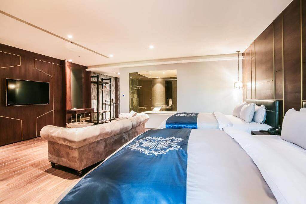【北港住宿】朝聖酒店的豪華套房內設有兩張大床，搭配深色木質牆面與藍色床旗，空間感寬敞明亮，並配有舒適的米色沙發與現代化視聽設備，展現高雅的居住氛圍。
