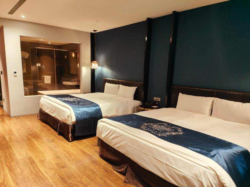 【北港住宿】朝聖酒店的雙人房設計以深藍色為主牆背景，床尾配有典雅的床旗，旁邊設有大型透明玻璃浴室，整體風格低調奢華且舒適。