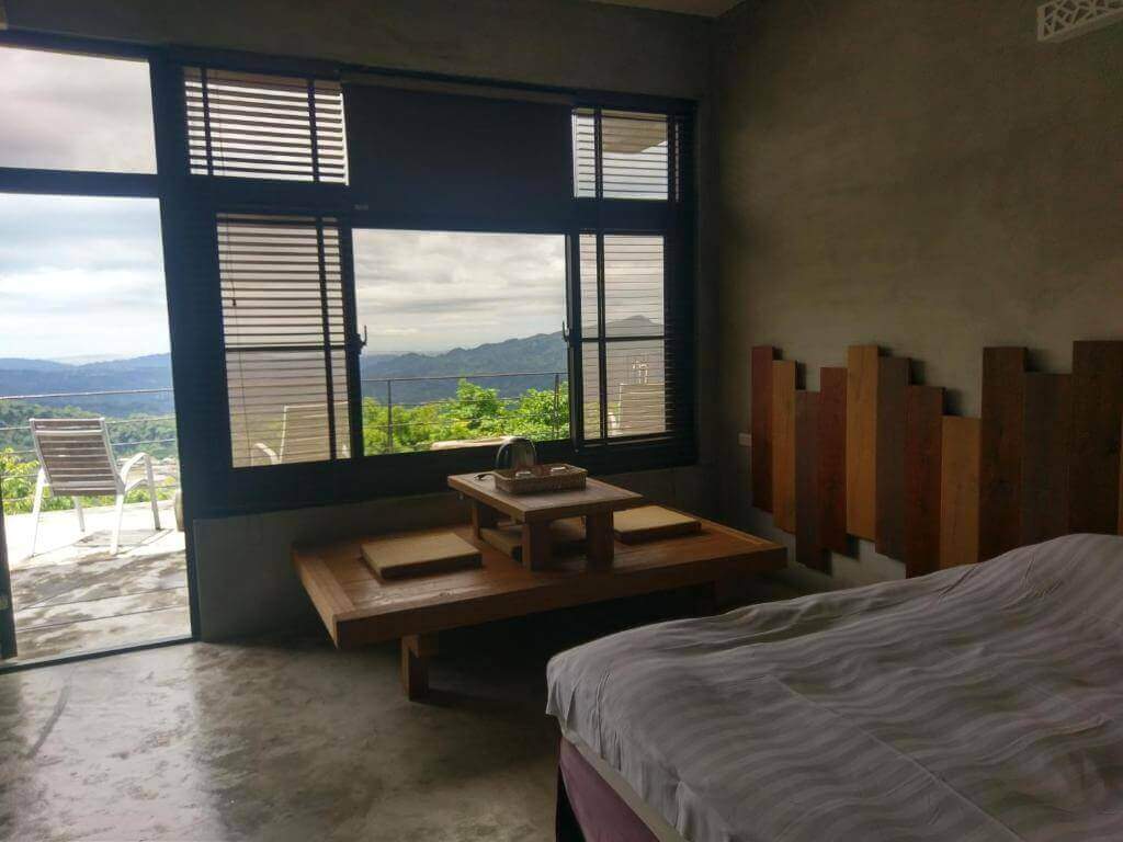 【大湖住宿】楓葉地圖房間內部設計簡約，牆面為灰色水泥質感，搭配木質裝飾床頭與矮式木桌。大面窗戶外可見山景與陽台椅，整體氛圍寧靜舒適，適合放鬆與欣賞自然景觀。