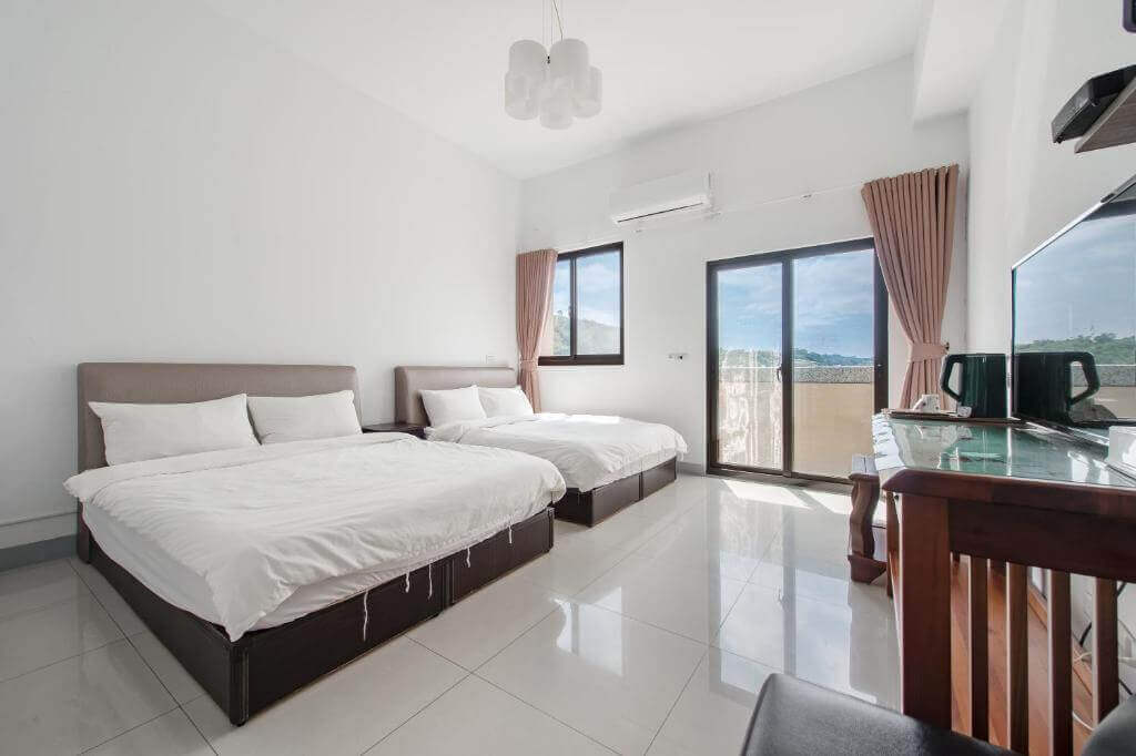 【大湖住宿】泉銘行館房間內設有兩張雙人床，白色牆面與淺色瓷磚地板增添空間感，陽台透光良好，窗簾柔和，搭配簡約的木質桌椅與基本家電，整體設計簡潔明亮。