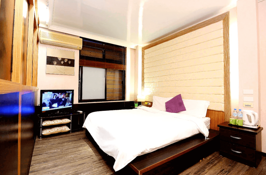 【烏來住宿】烏來名湯溫泉會館的房間內設有一張大床，背景牆以柔和的奶油色調設計，搭配紫色抱枕，增添色彩活力。窗外提供自然光線，室內配置小型電視、茶壺及飲用水，實用性強。