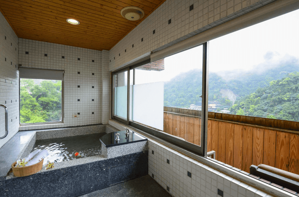 【烏來住宿】烏來名湯溫泉會館的浴室內設有一個帶窗的私人溫泉池，牆面使用白色方格瓷磚，屋頂以木質天花板設計，增添自然氛圍。大窗戶提供山景與自然光，讓泡湯更添愜意。