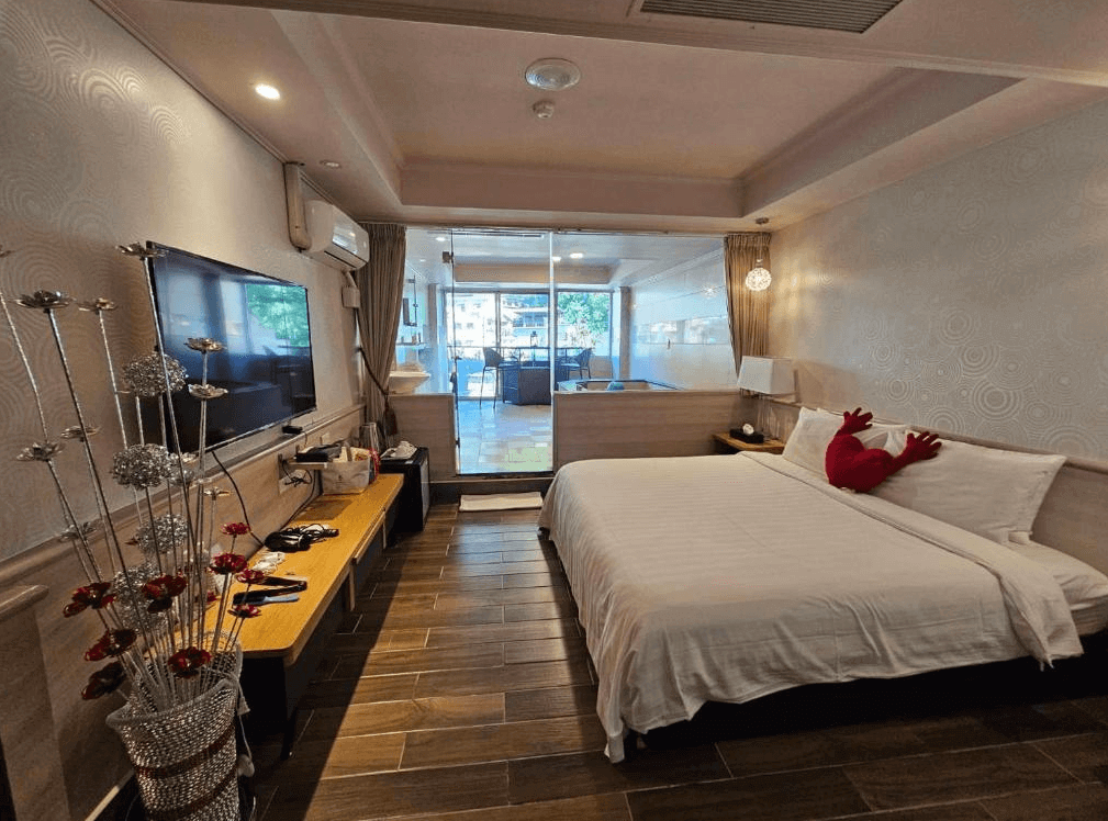 【烏來住宿】烏來水岸溫泉的房間內設有一張大床，床頭牆壁簡約而有層次感，配有溫馨的燈光照明。床尾設有平面電視與木製長桌，窗外可以看到自然景致，整體空間採光良好，給人舒適放鬆的氛圍。