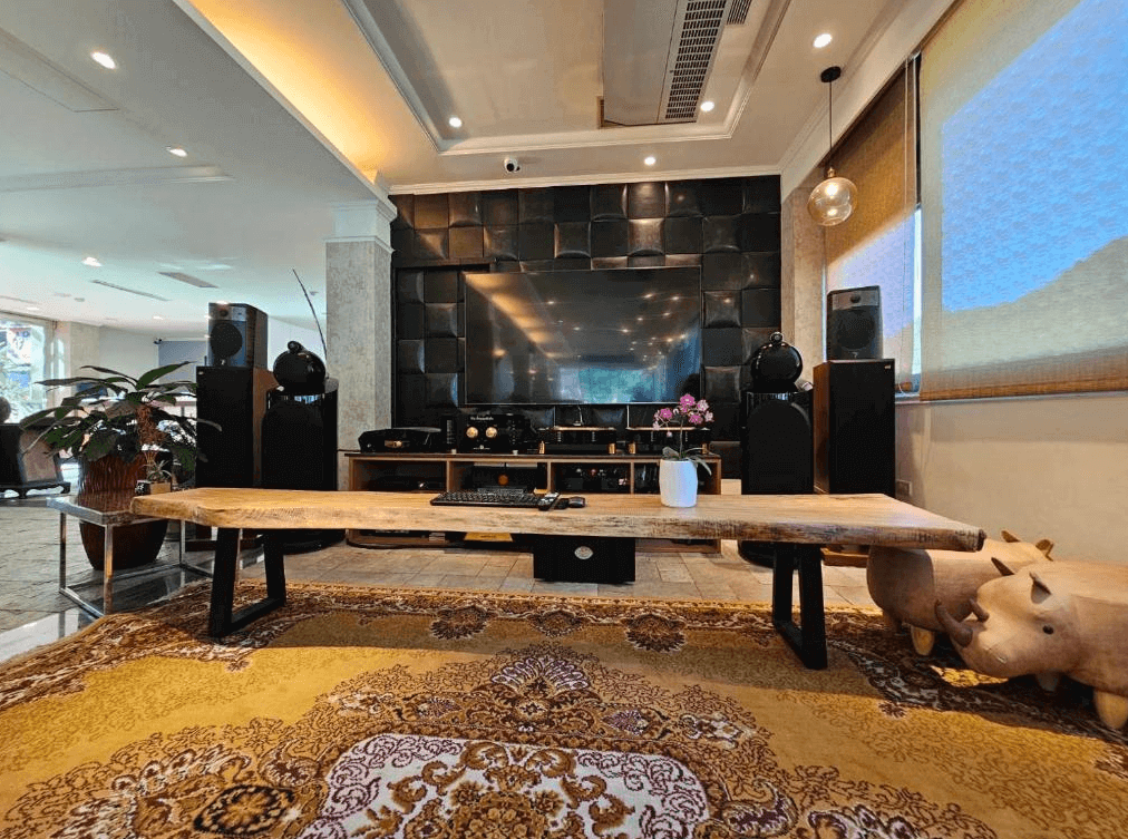 【烏來住宿】烏來水岸溫泉的客廳擺放了一張木質長桌，前方為高級影音設備，配有黑色音響與大屏幕電視，牆面採用深色設計，增添視覺層次感，營造豪華且沉穩的氛圍。