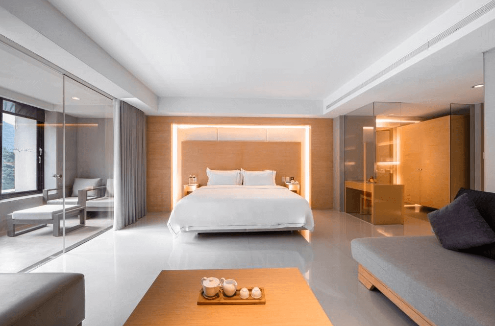 【烏來住宿】璞石麗緻溫泉會館的客房設計以簡約現代風格為主，採用白色與淺木色搭配，中央擺放一張大床，背景牆有隱藏式燈光。房間一側設有玻璃隔間，內含私人陽台與座椅，整體空間明亮且寧靜。