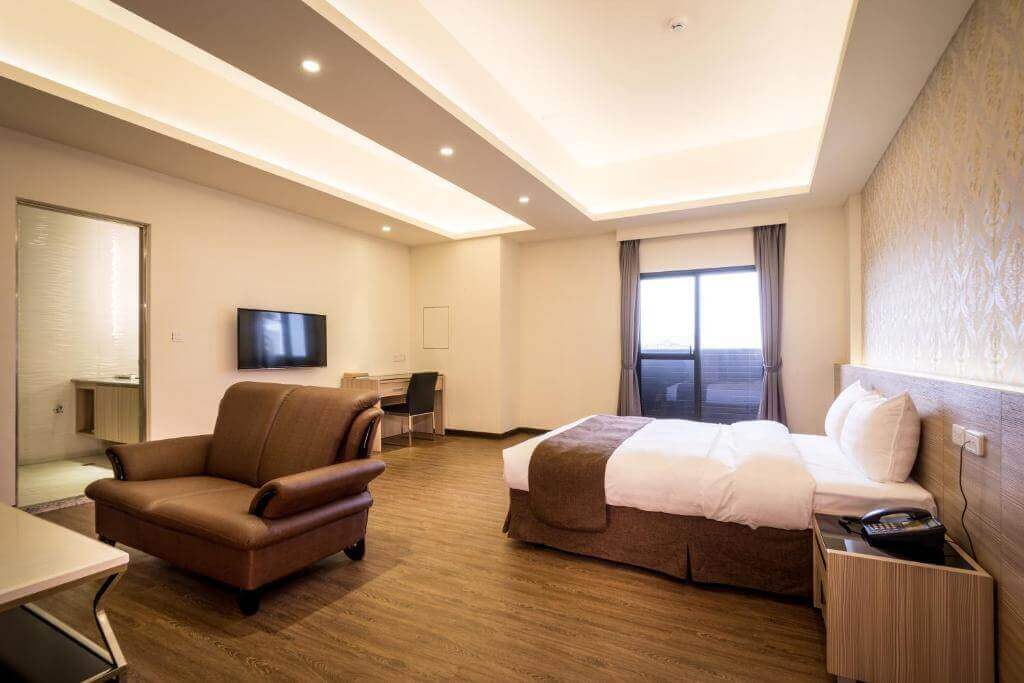 【北港住宿】禾都商務飯店的房間寬敞明亮，配有一張大床與棕色雙人沙發，窗外有陽台設計，搭配木質地板與簡潔的天花板燈光設計，整體風格溫馨而高雅。