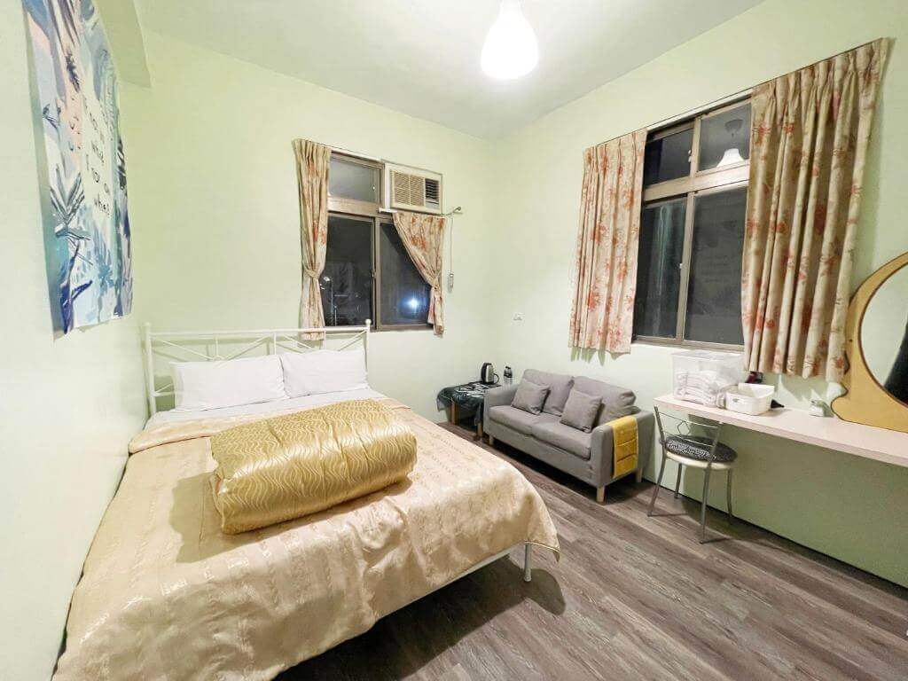 【美濃住宿】美濃德旺山莊的房間內景，淺綠色牆壁，擺放著一張雙人床，床上鋪有金黃色的被褥。旁邊有灰色雙人沙發和化妝台，房間採光良好，整體佈置簡單而溫馨。