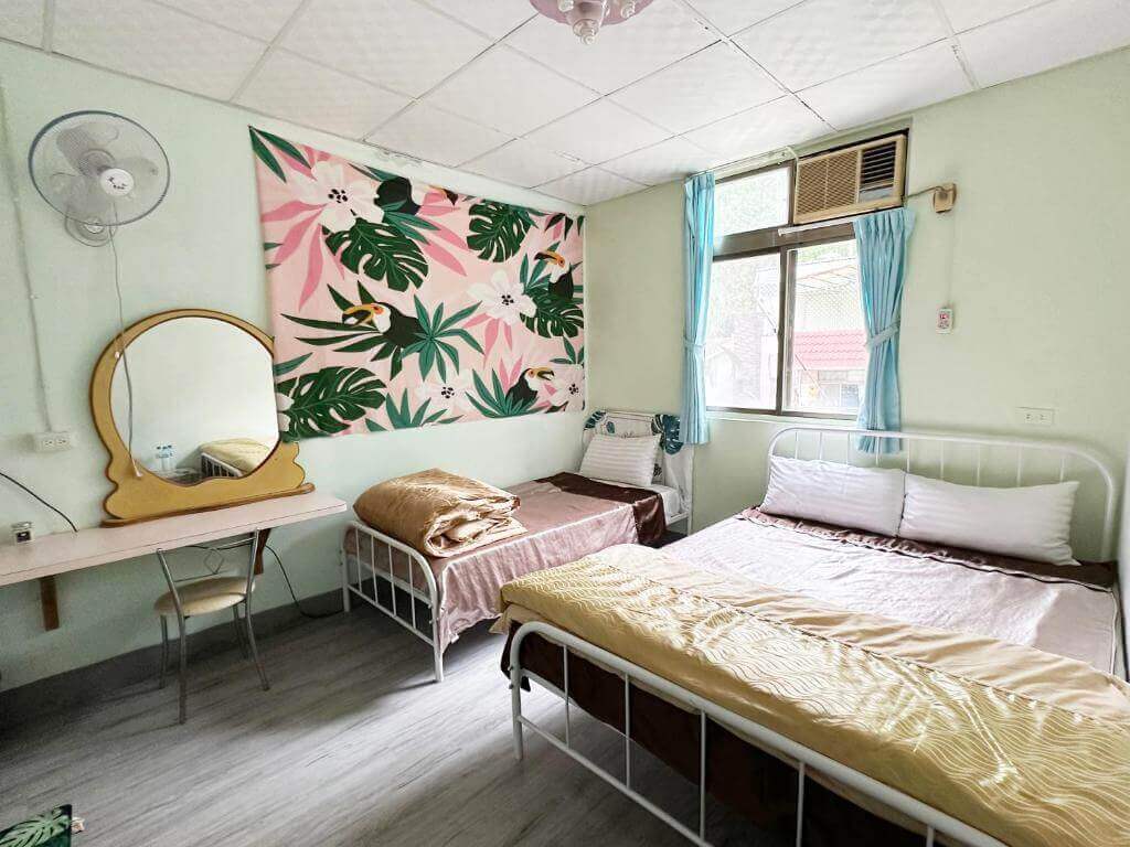 【美濃住宿】美濃德旺山莊的家庭房內景，牆上掛有熱帶植物和鳥類圖案的裝飾畫。房內有一張雙人床和一張單人床，牆壁為淡綠色，窗簾為天藍色，空間寬敞明亮，適合家庭入住。