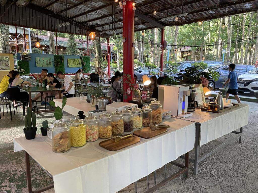 【美濃住宿】美濃德旺山莊的戶外用餐區，設有長桌和椅子，桌上擺放著多種零食和飲料，提供自助式餐點。背景為綠樹環繞的自然環境，氣氛輕鬆愉快。