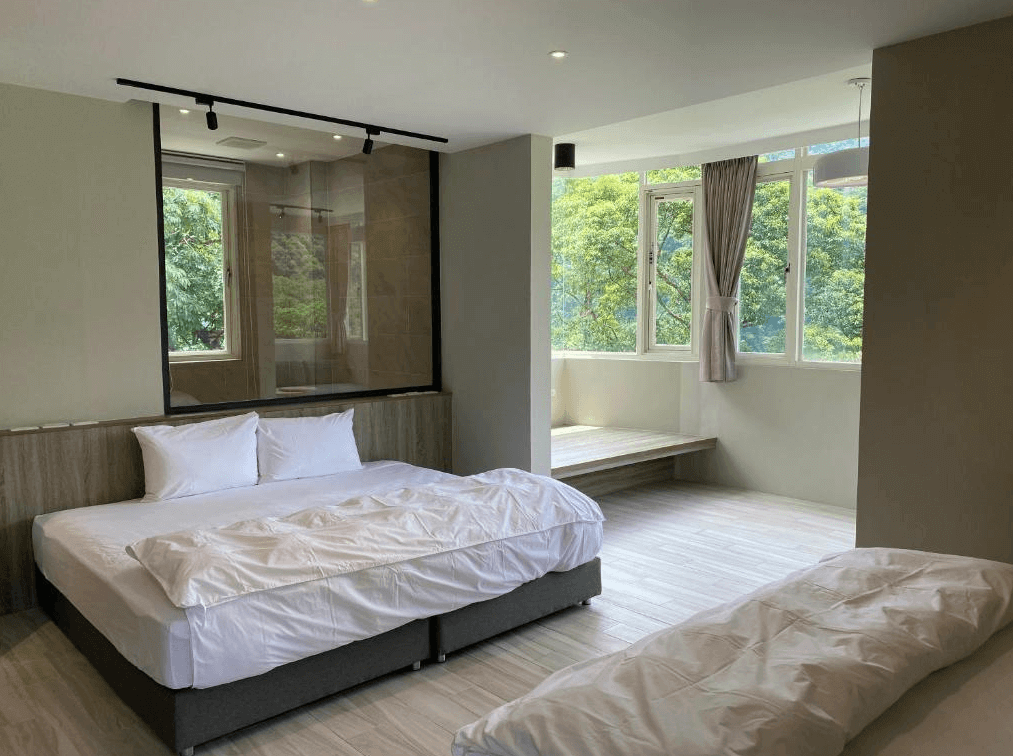 【烏來住宿】足立司拉 SLA ULAY的雙人房，設有大面窗戶，窗外可見綠意盎然的自然景觀。房間內有兩張大床，床頭背景牆使用簡約的木質設計，臥室與浴室之間採用透明玻璃隔間，增添視覺開闊感。