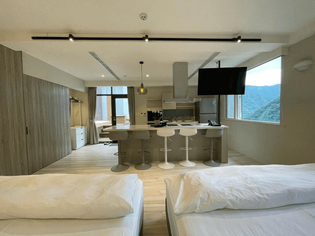 【烏來住宿】足立司拉 SLA ULAY的現代化客廳空間，配有吧檯椅的中島式設計，電視懸掛於空中，可靈活調整視角。大面窗戶讓自然光線灑入，背後可見群山環繞的美景。