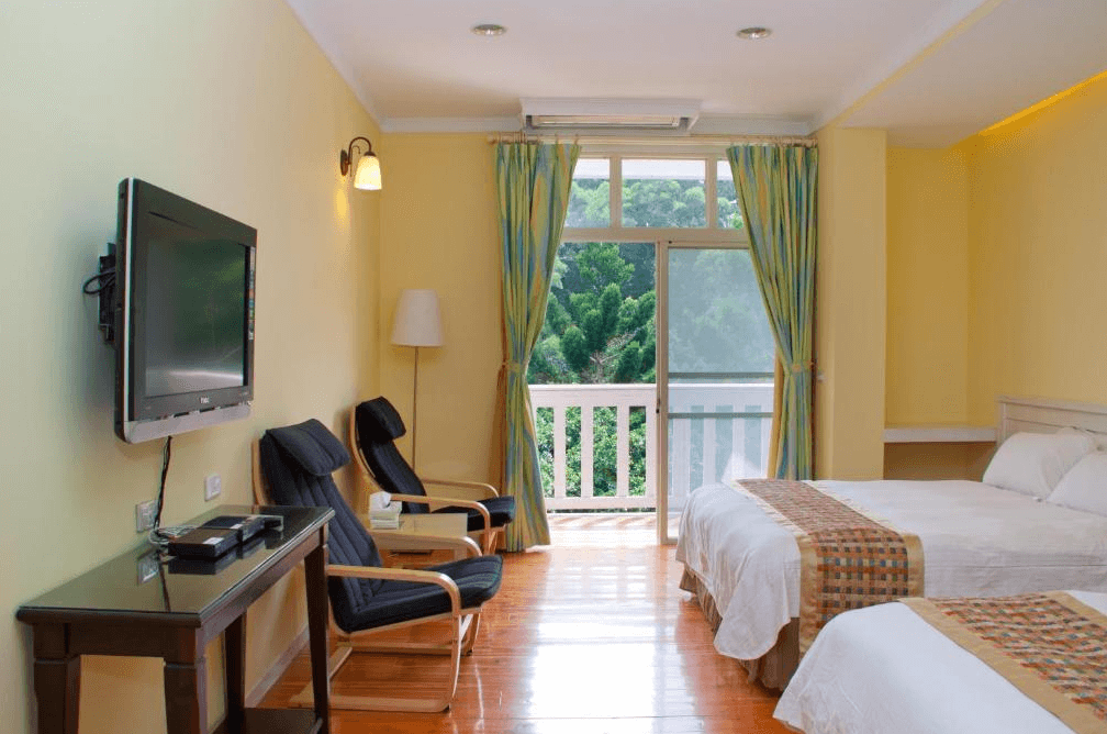 【通霄住宿】飛牛牧場充滿自然光的雙床客房，淺黃色牆面搭配藍綠色窗簾，明亮且舒適。陽台外是綠意盎然的樹林景色，室內設有兩張單人床、沙發區和電視，提供放鬆的住宿體驗。