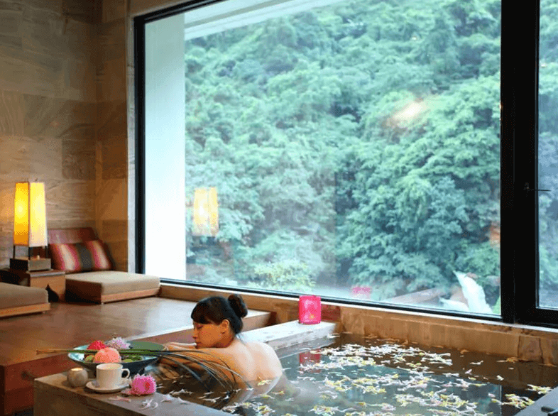 【烏來住宿】馥蘭朵烏來渡假酒店的私人溫泉池設於房間內，浴池周圍以木質和石材設計，窗外為翠綠山林景致，泡湯區擺設精緻花飾和茶杯，氛圍悠閒放鬆。