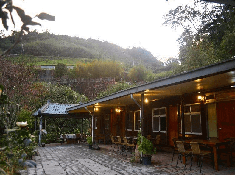 【阿里山住宿】仲明居陶藝坊的住宿外觀以簡樸的平房設計為主，周圍環繞著青山與花園景致，提供悠閒的戶外空間。庭院內設有多張桌椅，適合用餐或聚會活動。