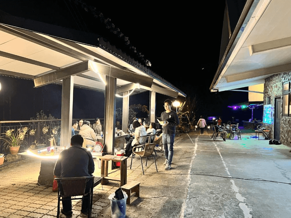 【阿里山住宿】仲明居陶藝坊夜晚的庭院被燈光點綴，溫馨而熱鬧。多組住客聚集在桌邊享受美食與交流，環境適合舉辦燒烤或晚宴活動。