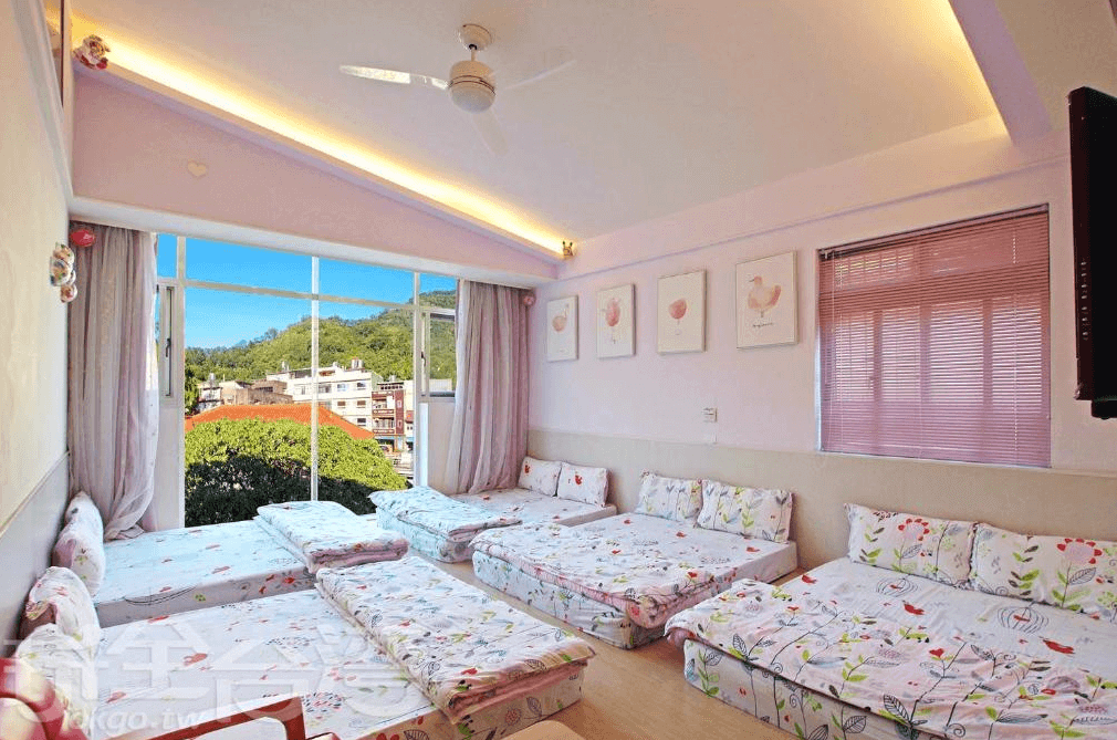 【司馬庫斯住宿】內灣-彎月民宿的家庭房，設有多組地墊床鋪，床單印有清新的花卉圖案，房間牆面為柔和的粉色，天花板裝有吊扇，大型落地窗外可見綠意盎然的山景與城鎮建築。