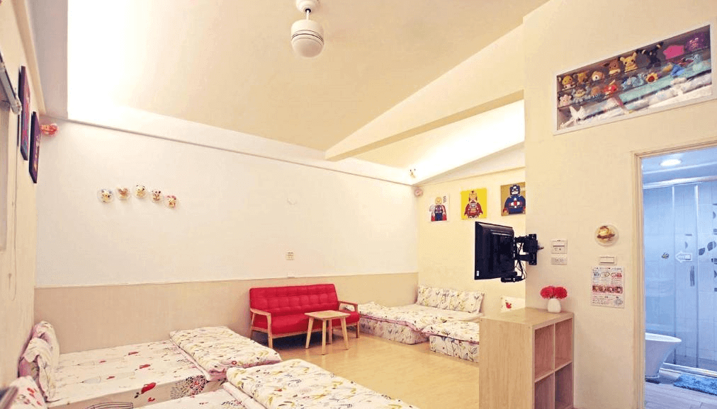 【司馬庫斯住宿】內灣-彎月民宿的多功能客廳，設有多組地墊床鋪和紅色沙發，牆面以簡單的白色為主，掛有多幅卡通裝飾畫，旁邊可見一間明亮的浴室，整體氛圍溫暖歡樂。