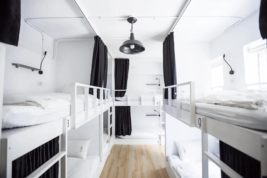 【台北車站住宿】【寧夏夜市住宿】"台北車站青年旅館"北門臥客青年旅舍的宿舍房，設有上下鋪床位，每張床位配有黑色窗簾提供隱私，整體以白色為主色調，搭配簡約的工業風吊燈，空間整潔明亮。