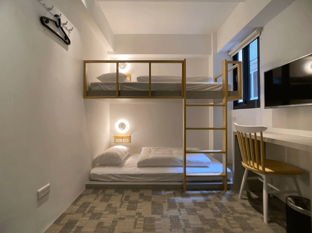 【台北車站住宿】【寧夏夜市住宿】"台北車站青年旅館"北門臥客青年旅舍上下鋪的客房，使用簡潔的木質設計，床鋪整潔，房內設有書桌、電視和掛衣架，窗戶提供自然採光，空間溫馨且實用。