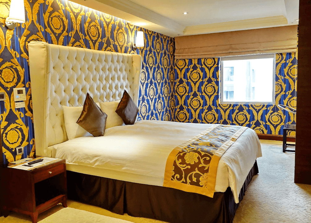 【寧夏夜市住宿】台北互舍酒店的豪華雙人房，設有帶軟墊的白色床頭板，牆面使用藍色和金色交織的華麗壁紙，房間內有寬敞的窗戶提供自然採光，裝潢富有皇家風格。