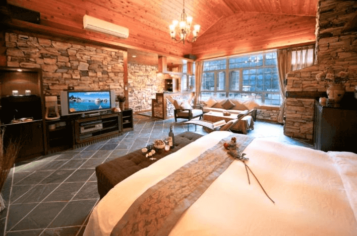 【廬山溫泉】名廬假期大飯店典雅奢華的鄉村風套房，採用大量原石與木質裝潢，搭配挑高木質天花板與水晶吊燈，營造出溫暖舒適的氛圍。房內設有加大雙人床、舒適沙發區與家庭娛樂設備，大片落地窗讓自然光線灑入，帶來寧靜放鬆的住宿體驗。