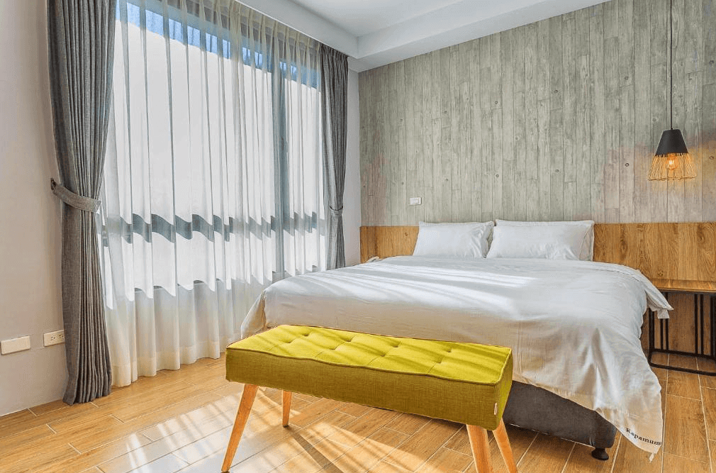 【日月潭住宿】嘎巴暮暮的雙人房設計簡潔，背景牆以木紋與水泥風混搭，展現現代與自然結合的美感。床前擺放一張亮黃色的長椅作為點綴，整體氛圍舒適且活潑。
